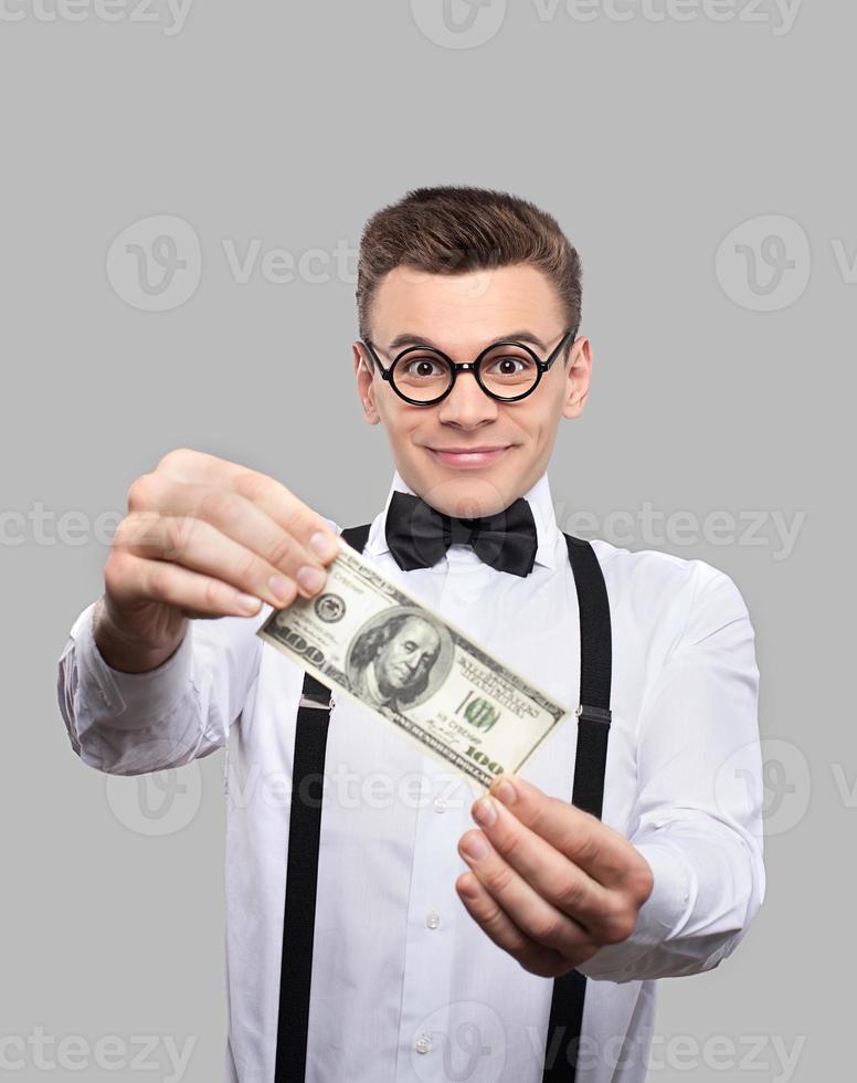 His first earnings. Cheerful young man in bow tie and suspenders holding a one hundred dollar bill and smiling while standing against grey background photo