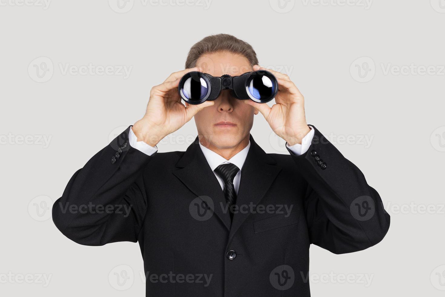 Looking for the new opportunities. Confident mature man in formalwear looking through binoculars while standing against grey background photo