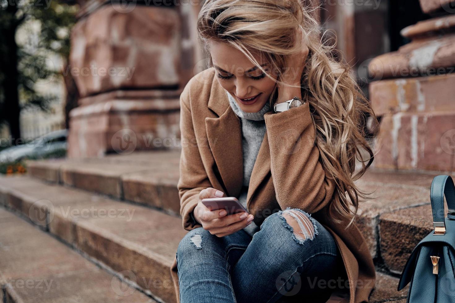 chatear con amigos, chateando con amigos. hermosa joven usando su teléfono inteligente y sonriendo mientras se sienta en las escaleras al aire libre foto