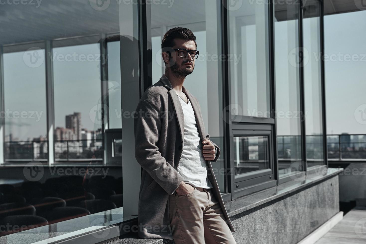 Young and successful.  Confident young man in eyewear keeping hand in pocket and looking away while leaning on the office building outdoors photo