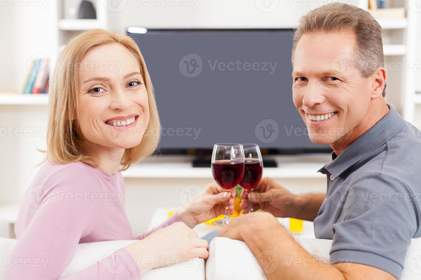 Happy to be together. Rear view of smiling mature couple sitting in front of TV and holding glasses with red wine photo