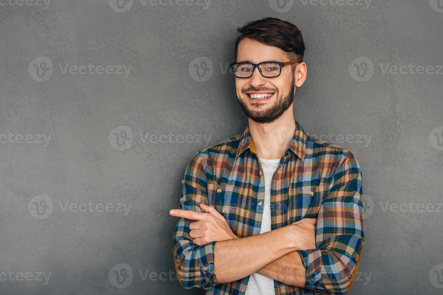 This is my choice Cheerful young man in glasses pointing at copy space and looking at camera with smile while standing against grey background photo