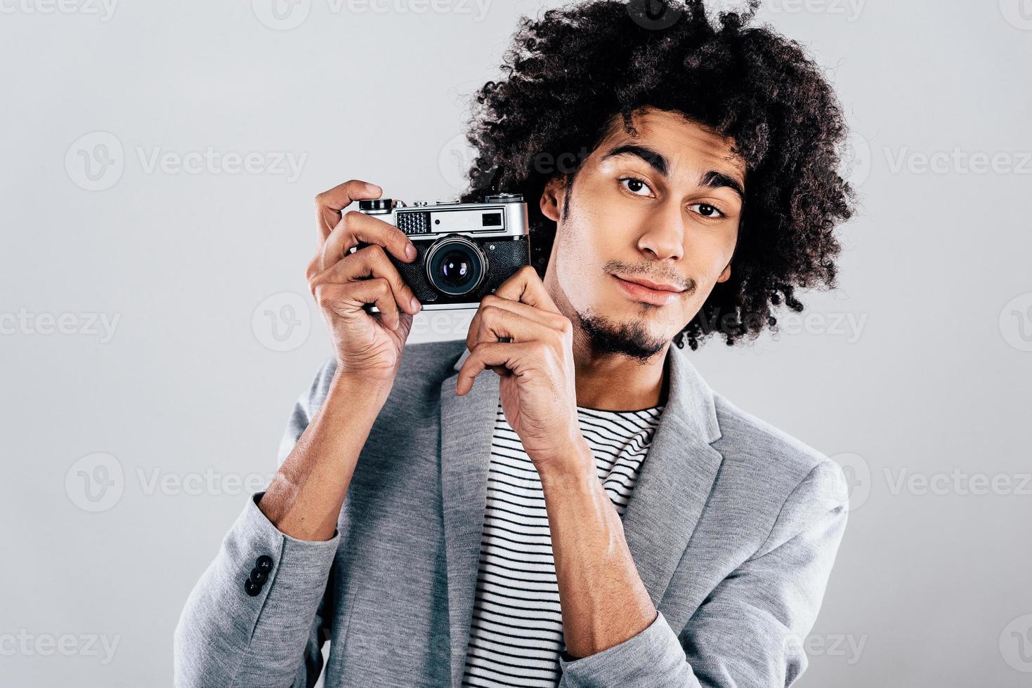 muéstrame tu mejor sonrisa guapo joven africano sosteniendo una cámara de estilo retro y mirando a la cámara mientras está de pie contra un fondo gris foto