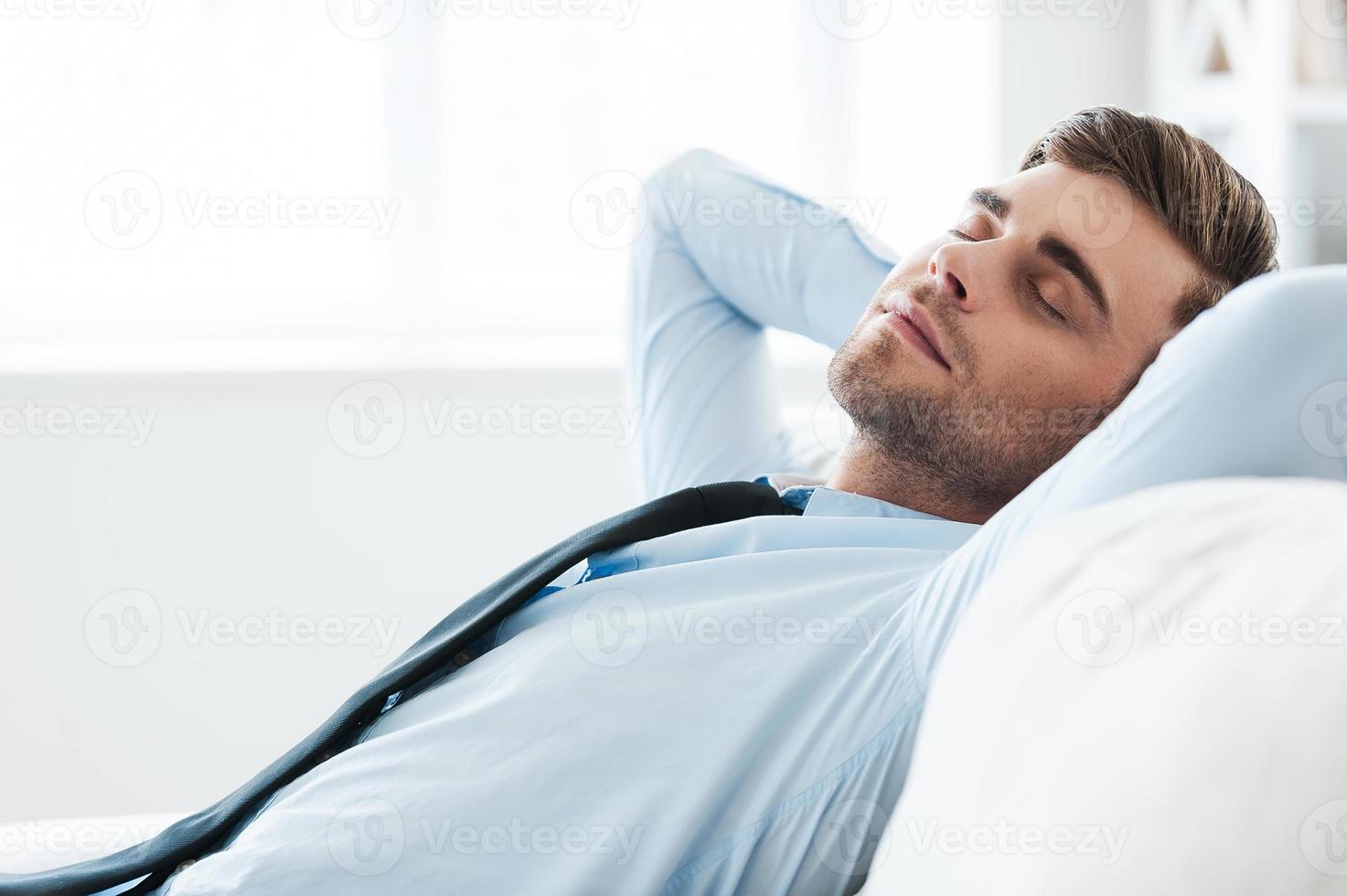 Time for relaxation. Handsome young businessman holding hands behind head and keeping eyes closed while sitting on sofa photo