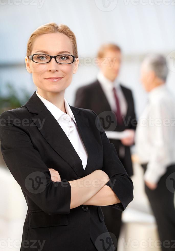 Confident in herself. Cheerful mature woman in formalwear keeping arms crossed and smiling while her colleagues talking on background photo