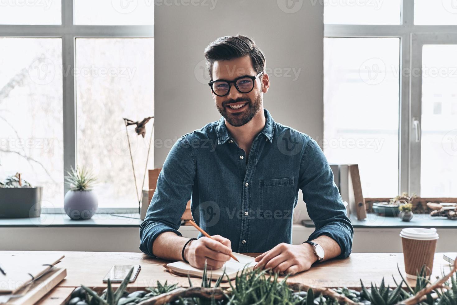 joven y exitoso. un joven apuesto escribiendo algo en un organizador personal y sonriendo mientras está sentado en la oficina foto