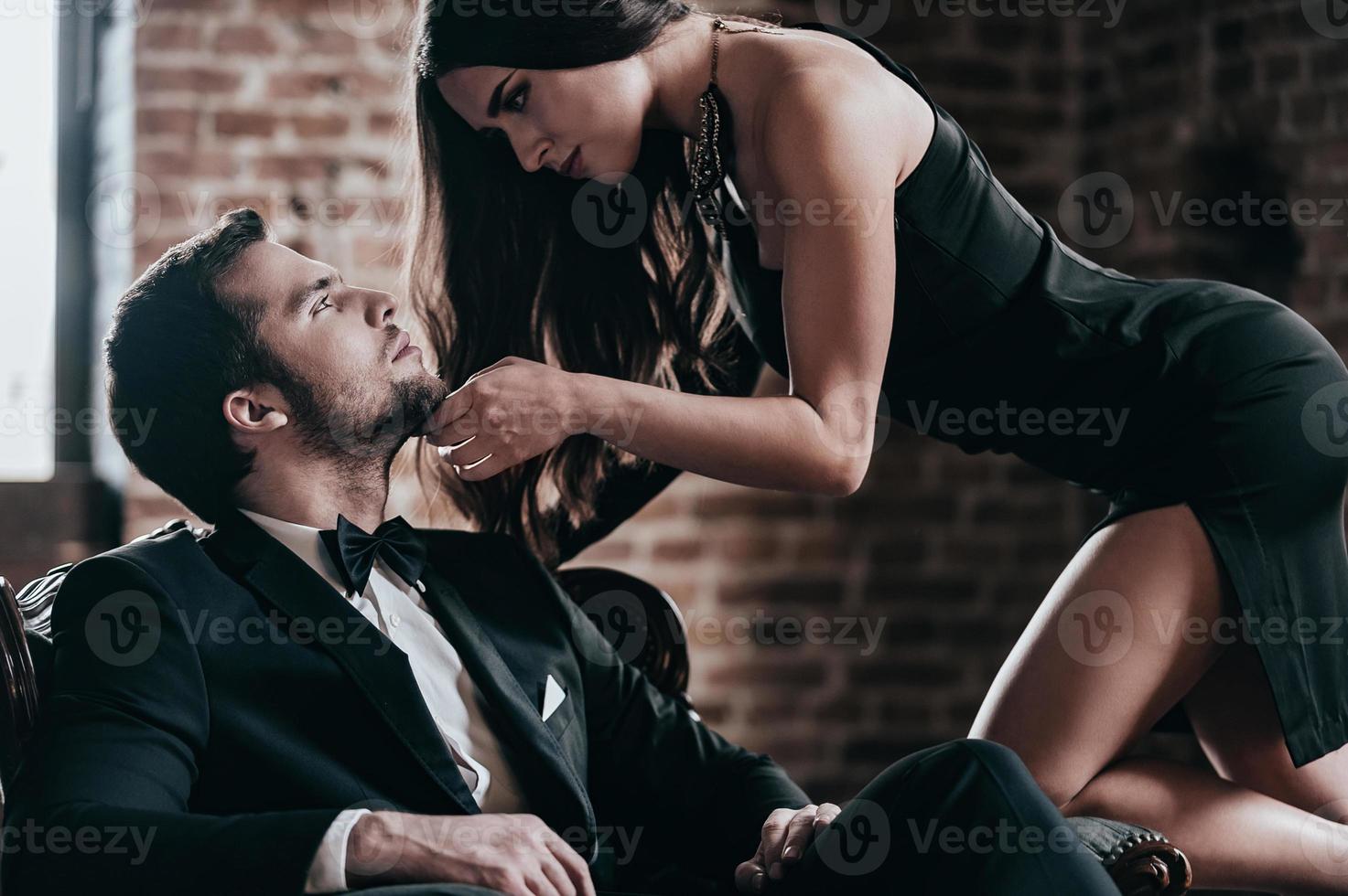 deseo desatado. bella joven vestida de cóctel inclinándose hacia su novio sentado en una silla mientras se miran en el interior del loft foto