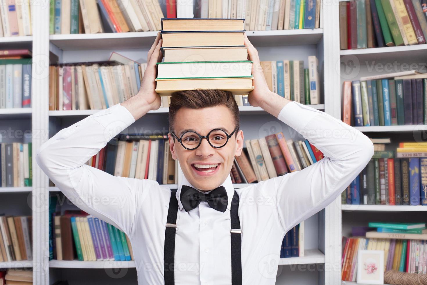 He loves studying. Cheerful young man in shirt and bow tie sitting at the table in library and holding a book stack on his head photo