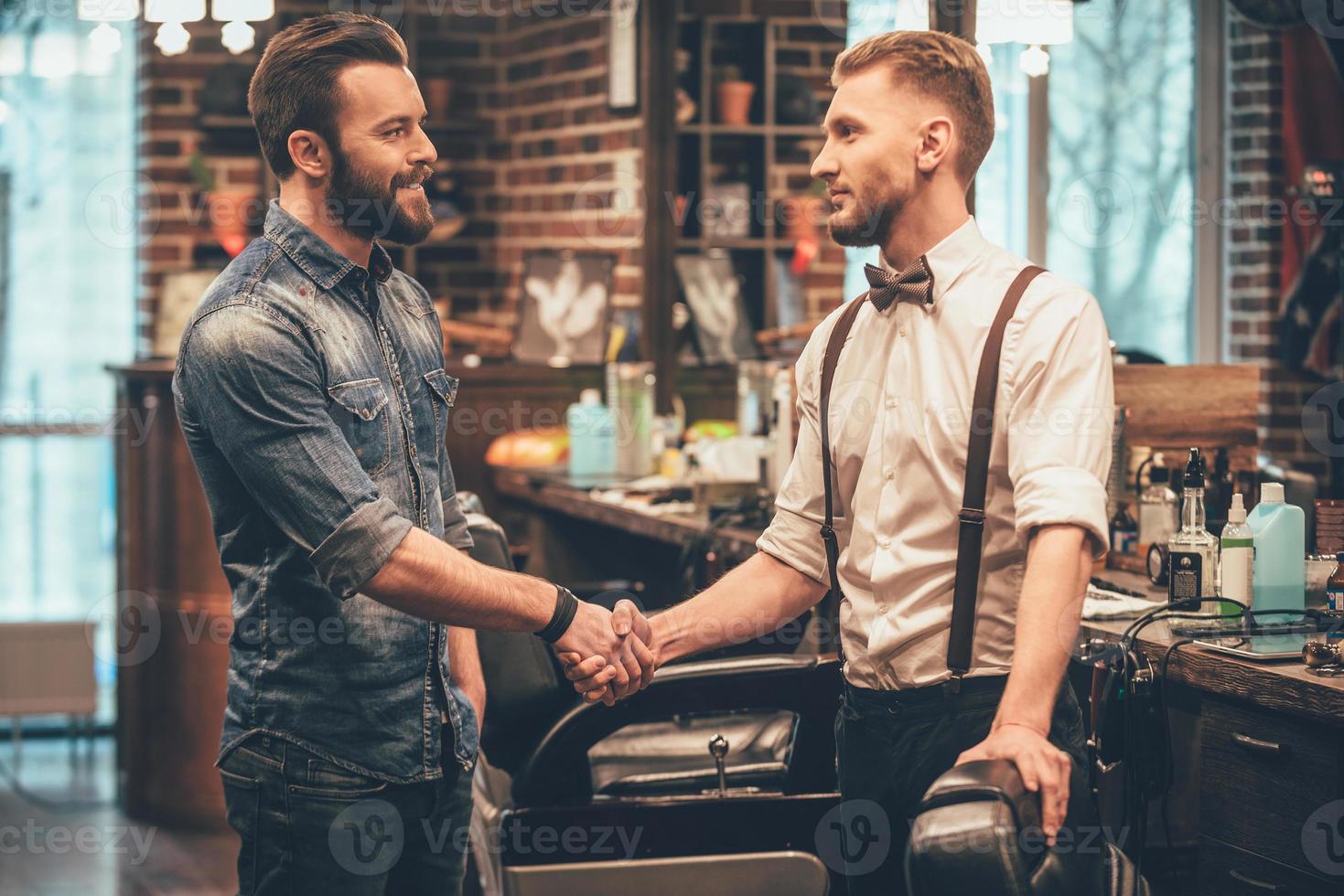 Thank you for quality haircut Barber and his client shaking hands with smile while standing at barbershop photo