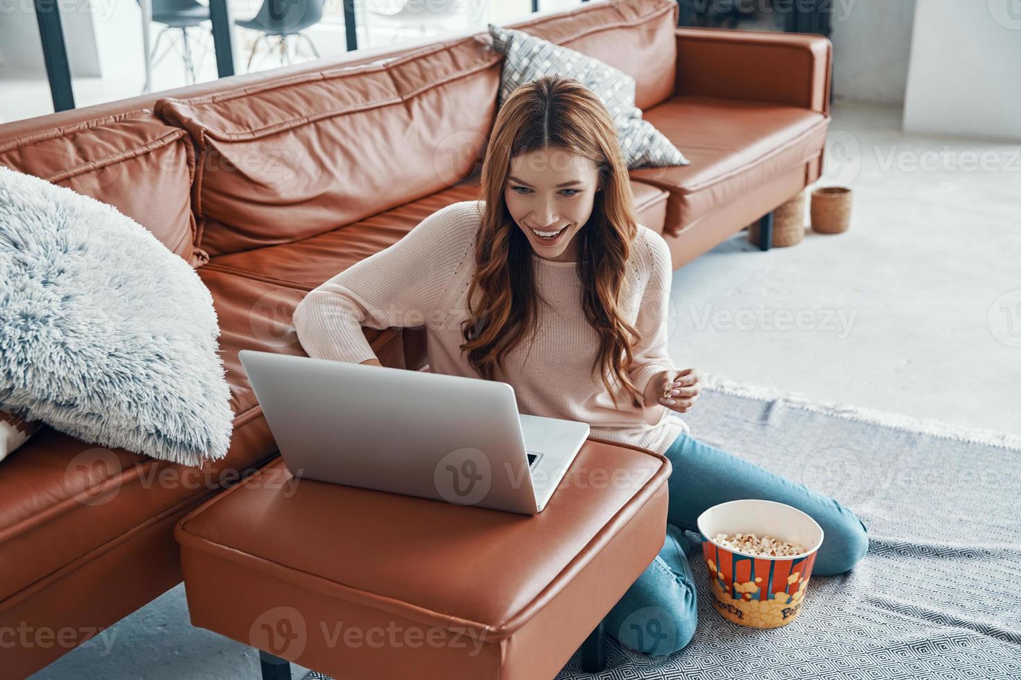 Attractive young woman using laptop and eating popcorn while spending time at home photo