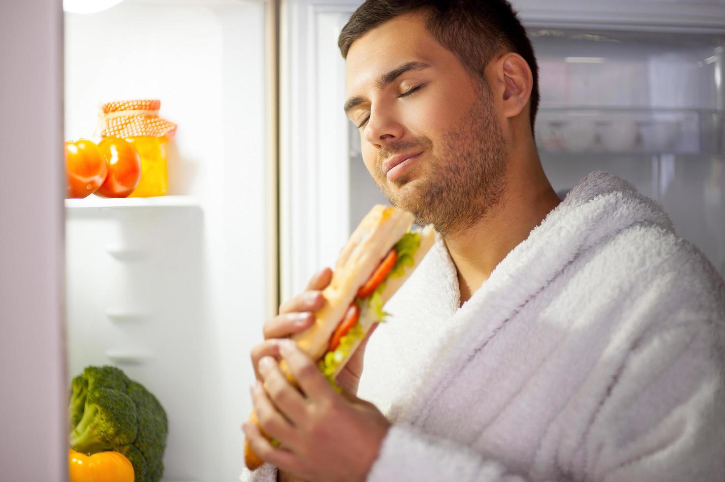 The best sandwich ever. Satisfied young man in bathrobe standing near the open fridge and eating sandwich with his eyes closed photo