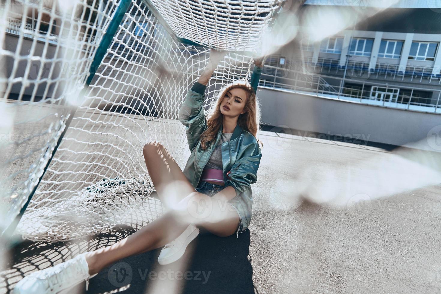 belleza de moda. mujer joven atractiva con ropa informal tocando el poste de la portería mientras se sienta al aire libre foto