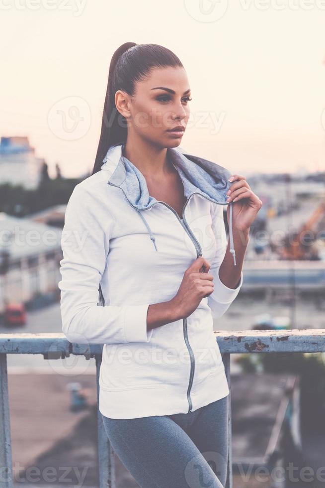 Ready for training. Beautiful young woman adjusting her sports clothes and looking away while standing on the bridge photo