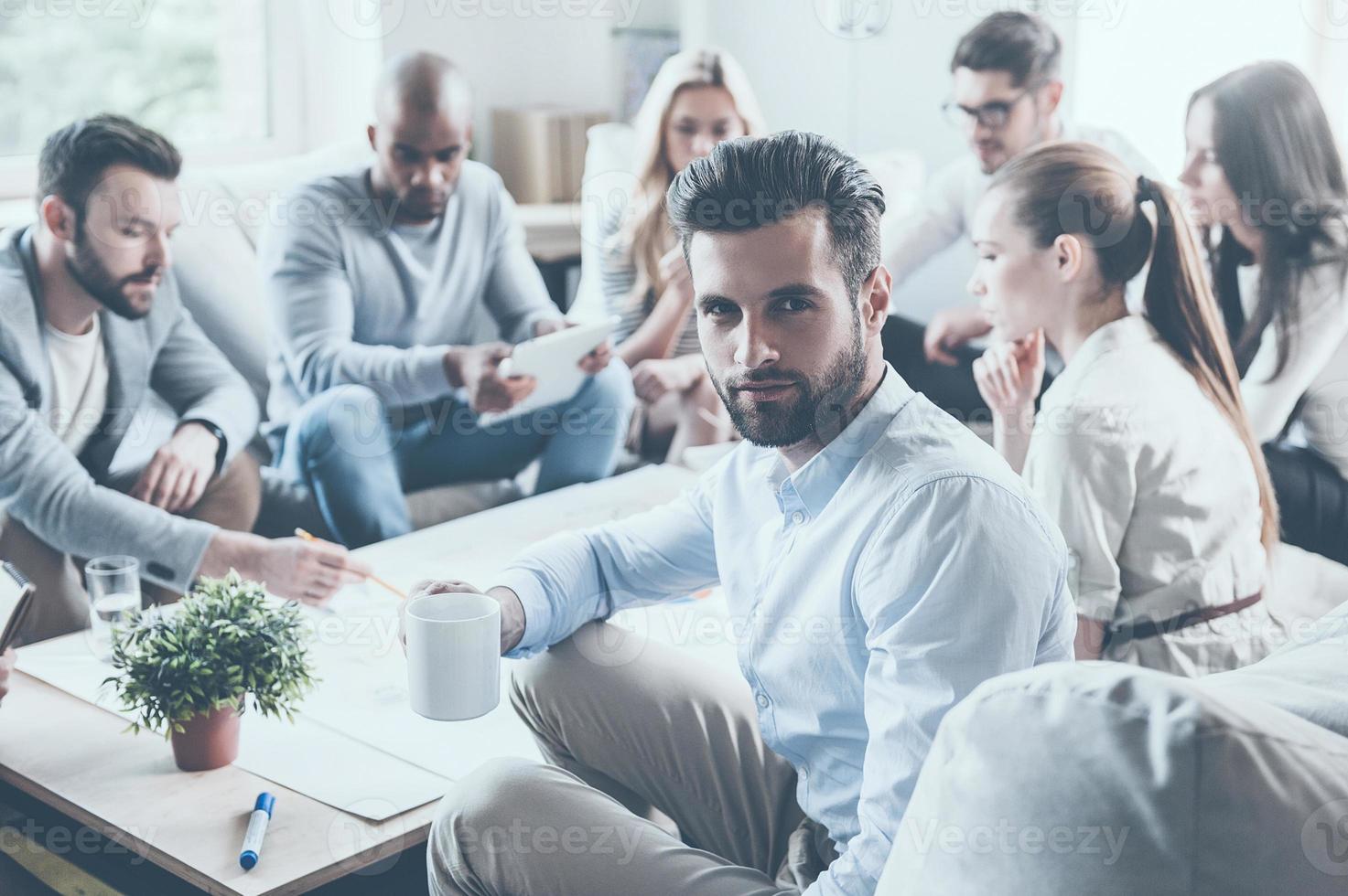 Feeling confident in my team. Group of confident business people sitting at the office desk together and discussing something while young man holding coffee cup and looking over shoulder photo