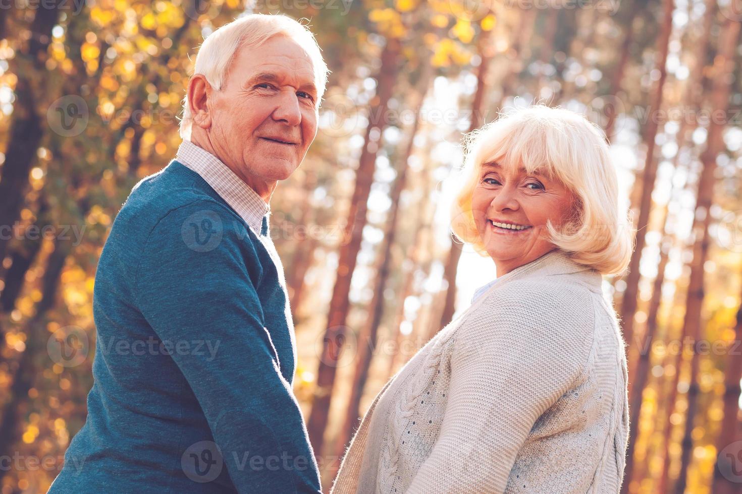 We love spending time together. Rear view of happy senior couple holding hands and looking over shoulder while walking by park together photo