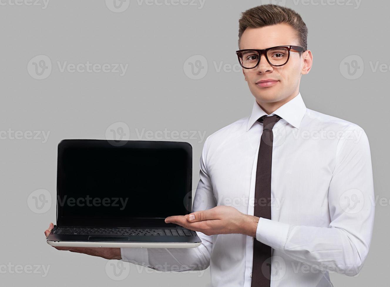 Man with laptop. Handsome young man in shirt and tie holding a laptop and pointing it while standing against grey background photo