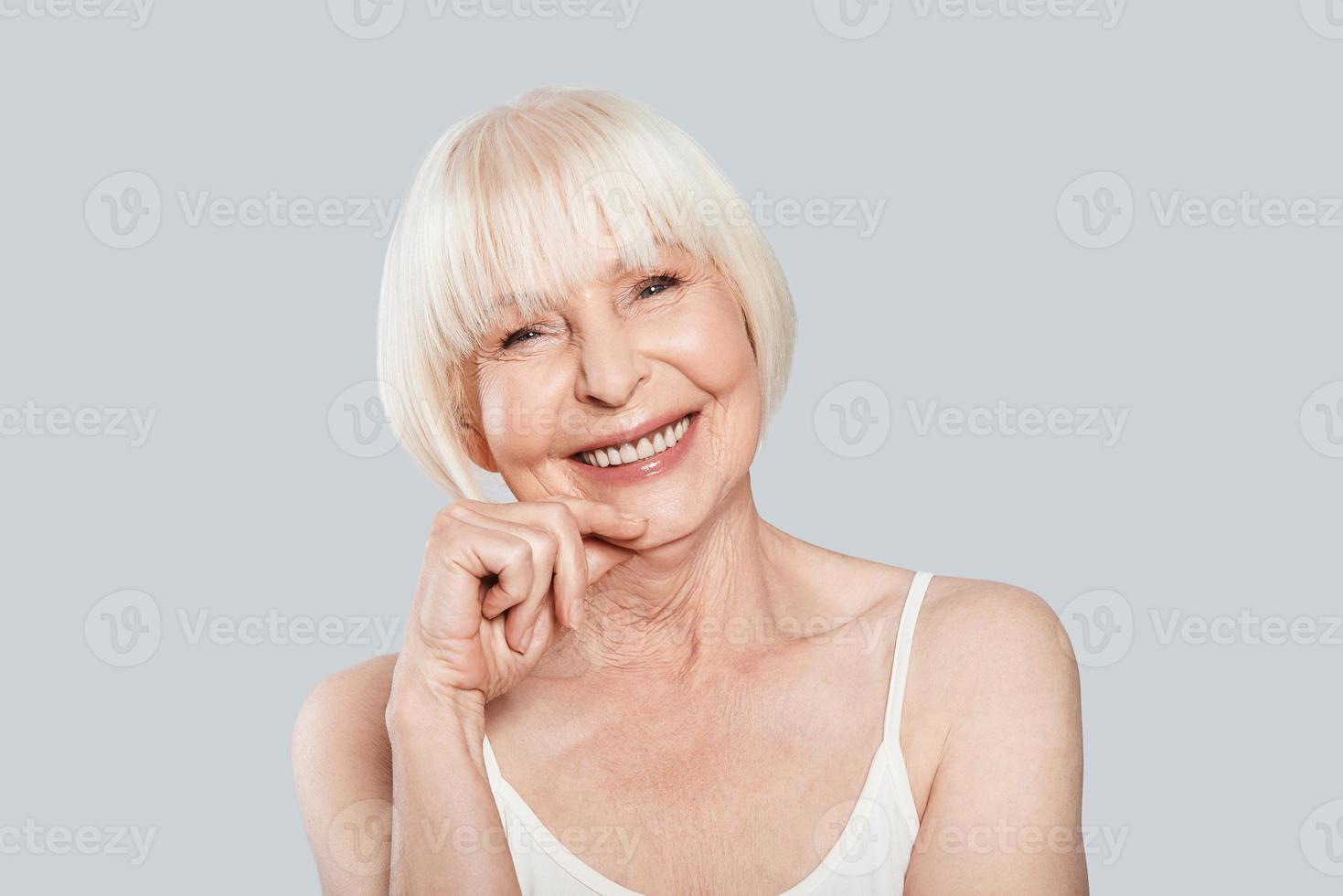 sonrisa sincera. hermosa anciana mirando a la cámara y sonriendo mientras está de pie contra el fondo gris foto