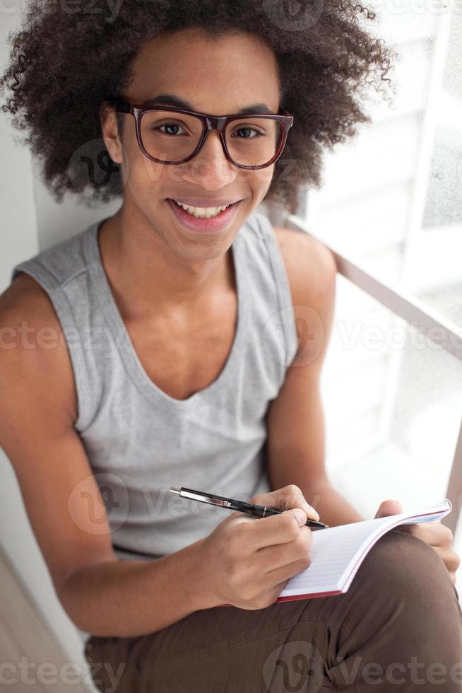 Just inspired. Top view of smiling teenage African boy sitting on the window sill and looking at camera while holding note pad in his hands photo