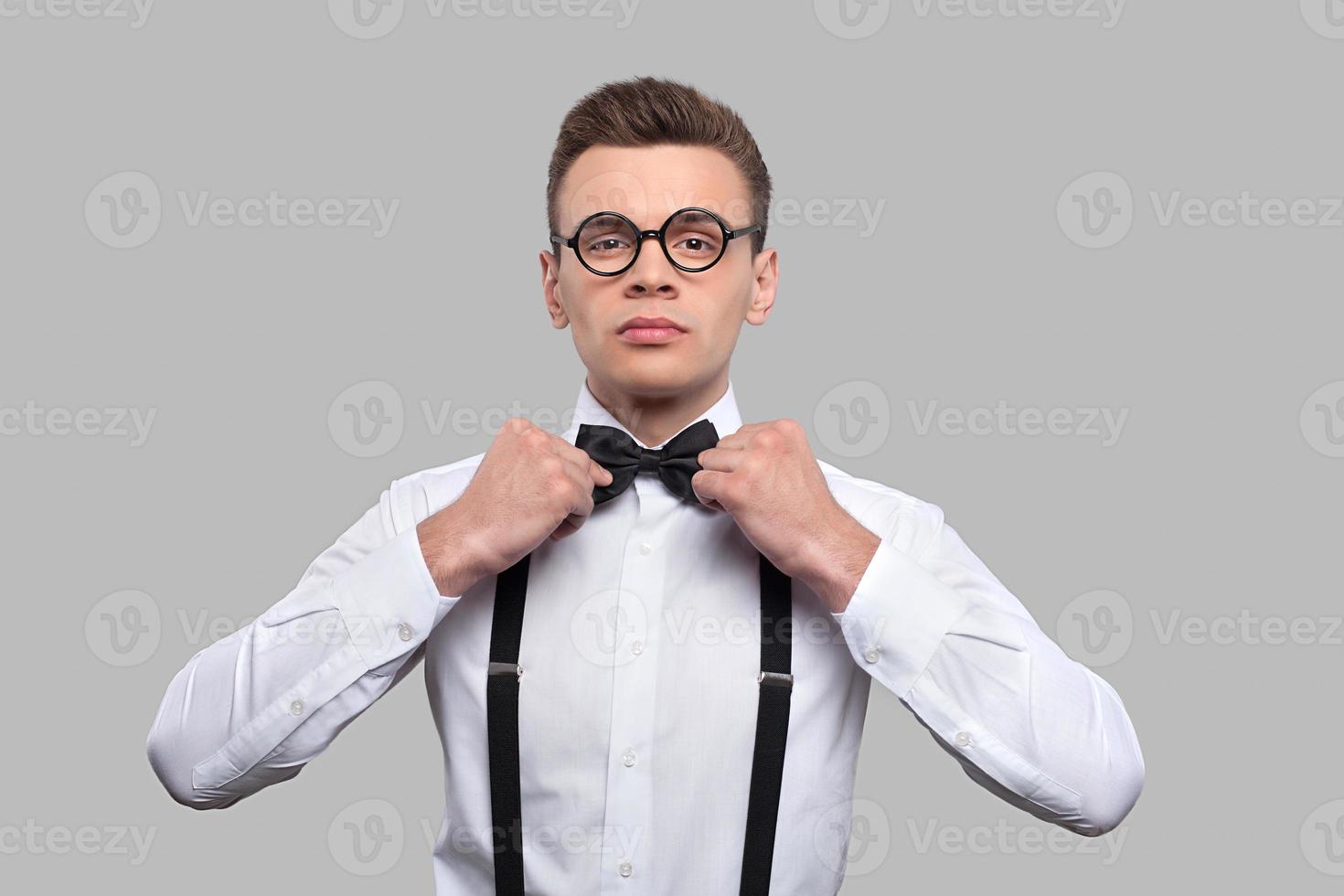 Confident in his skills. Serious young nerd man adjusting his bow tie and looking at camera while standing against grey background photo