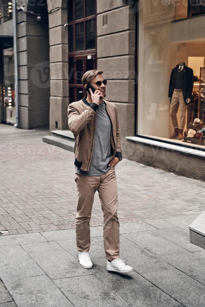 Amazing day in the city. Full length of handsome young man in casual wear talking on his smart phone and smiling while walking through the city street photo