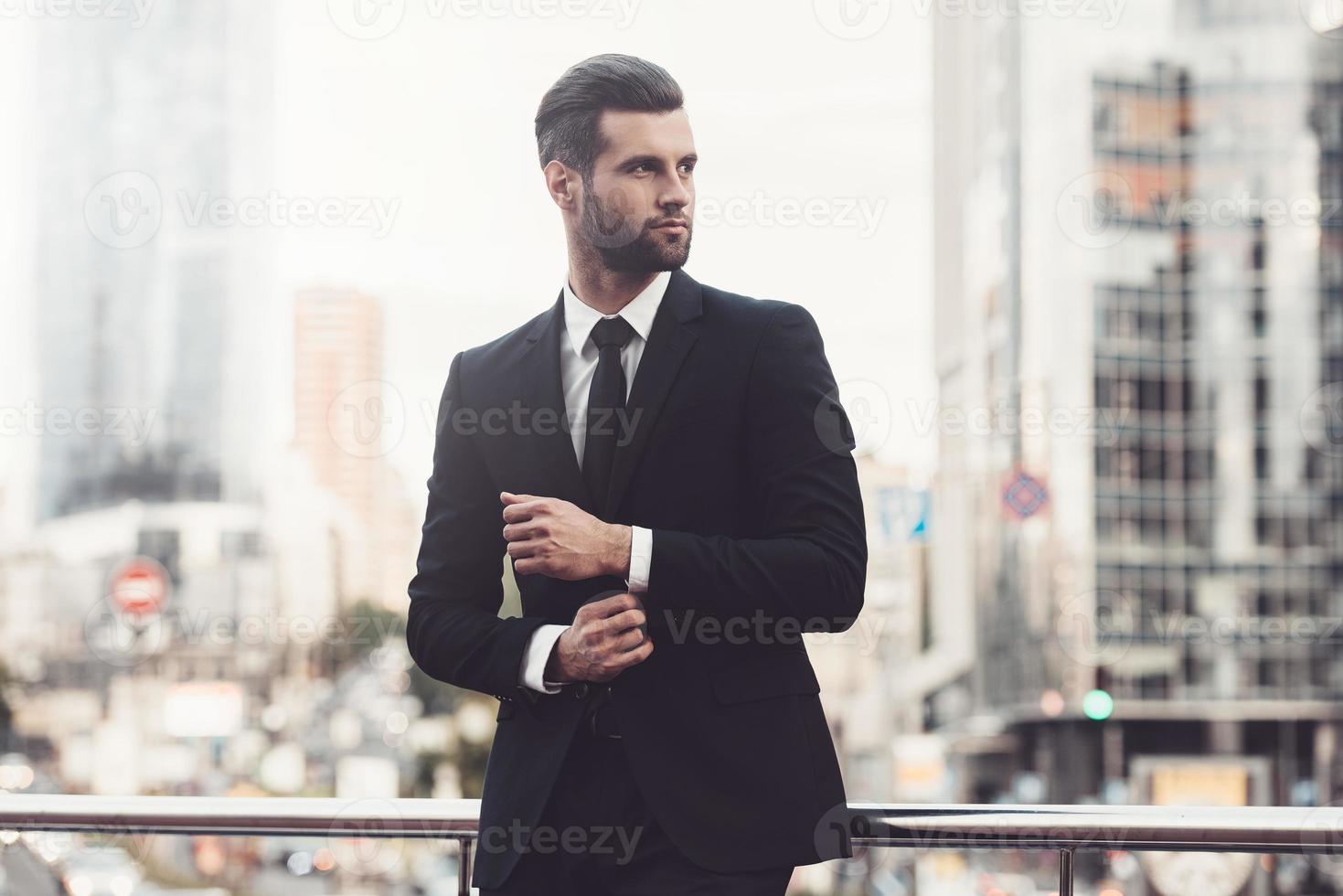 hombre de negocios moderno. joven confiado con traje completo ajustando su manga y mirando hacia otro lado mientras está de pie al aire libre con el paisaje urbano en el fondo foto