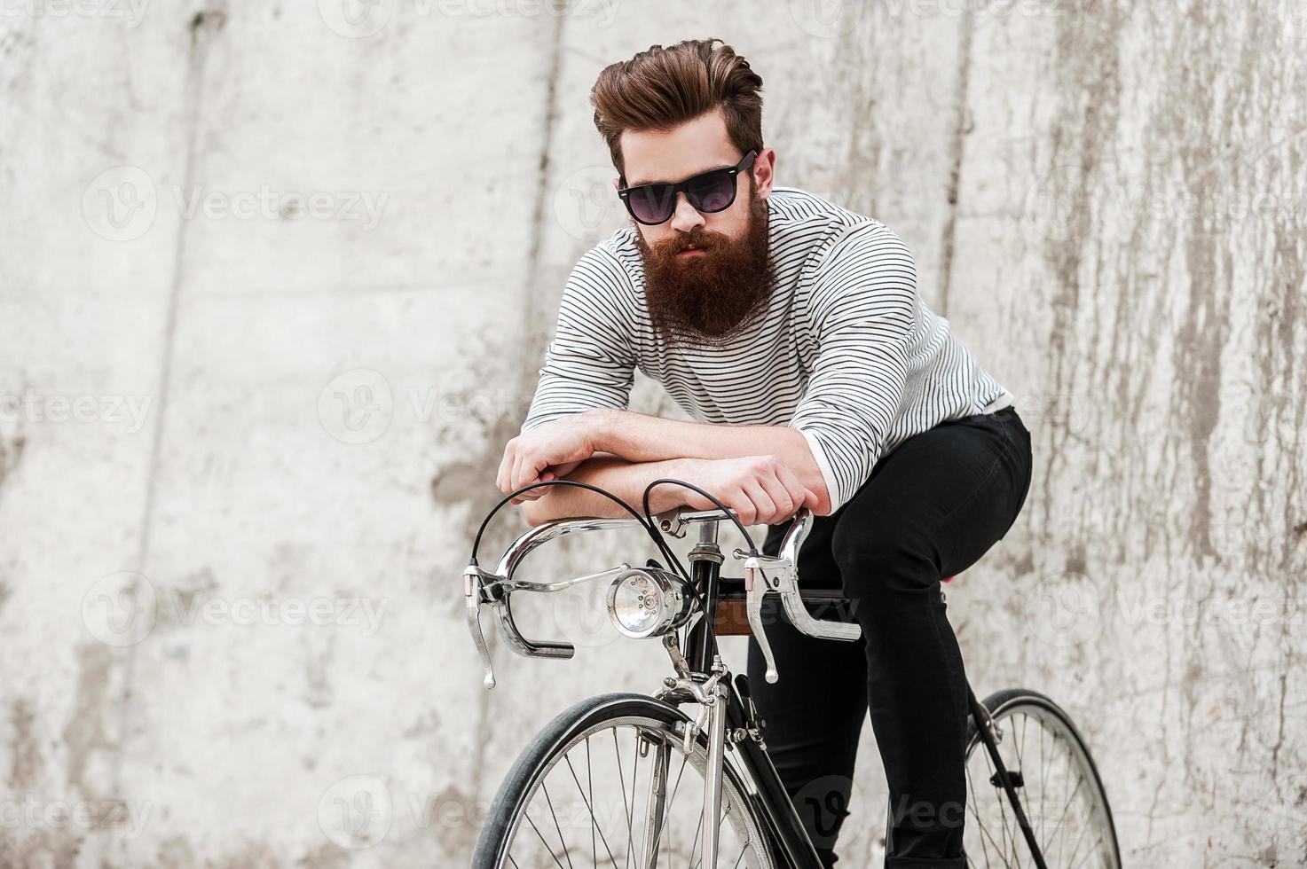 Choosing green transport. Handsome young bearded man leaning at his bicycle while sitting on it against the concrete wall photo
