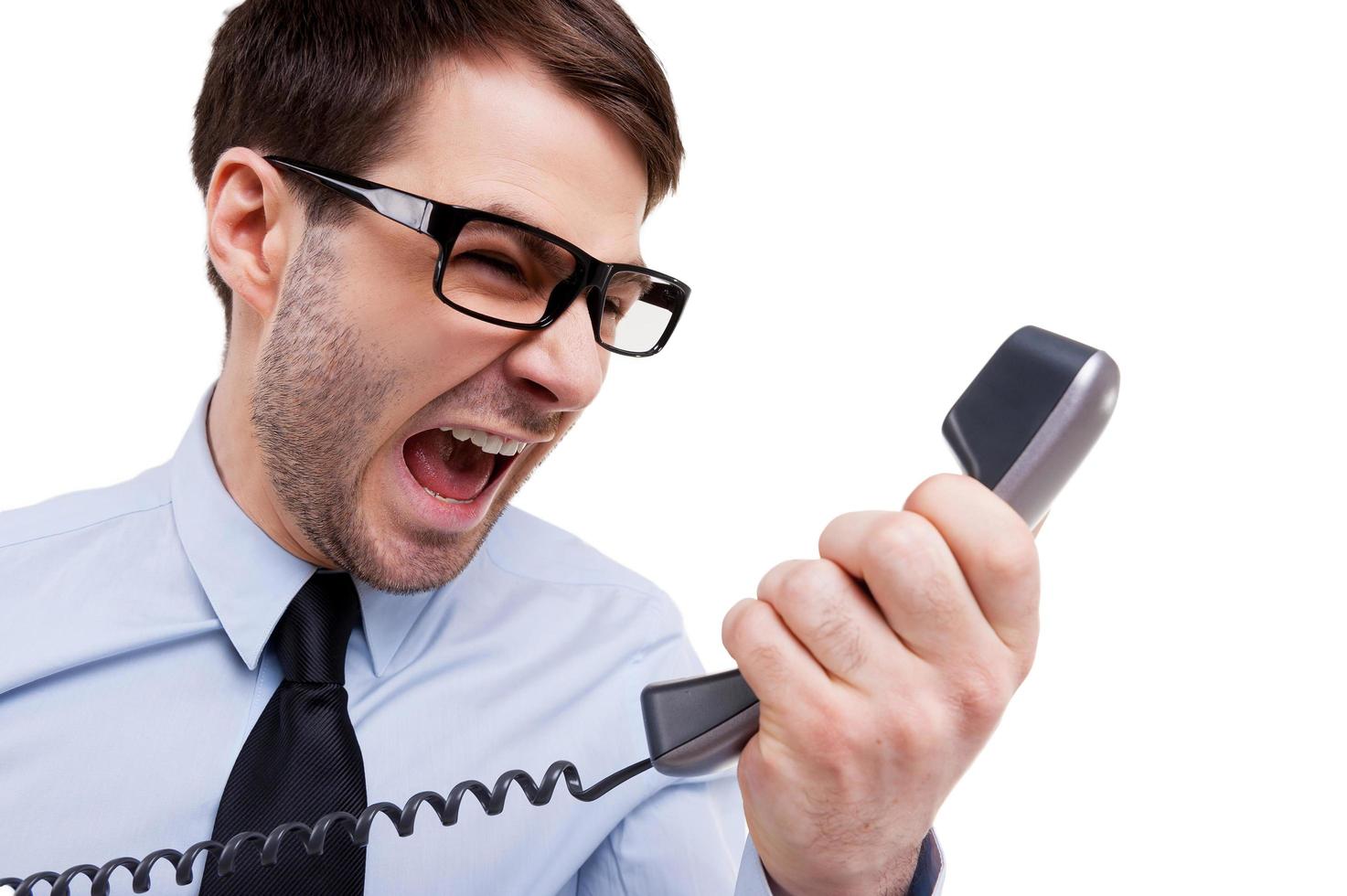 Stressful office life. Furious young man in shirt and tie shouting at telephone while standing isolated on white photo