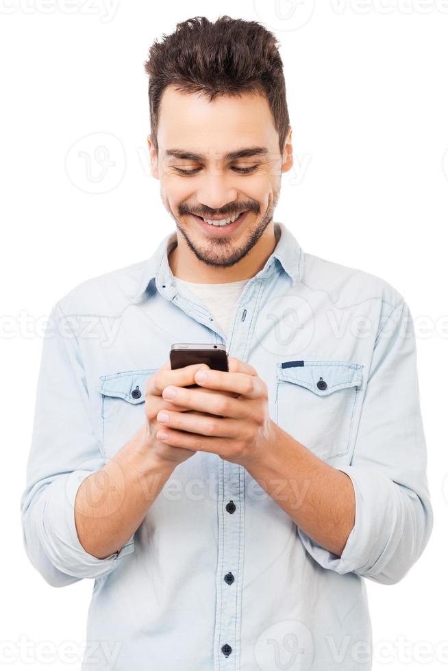 Nowhere without my phone. Handsome young man holding mobile phone and smiling while standing against white background photo