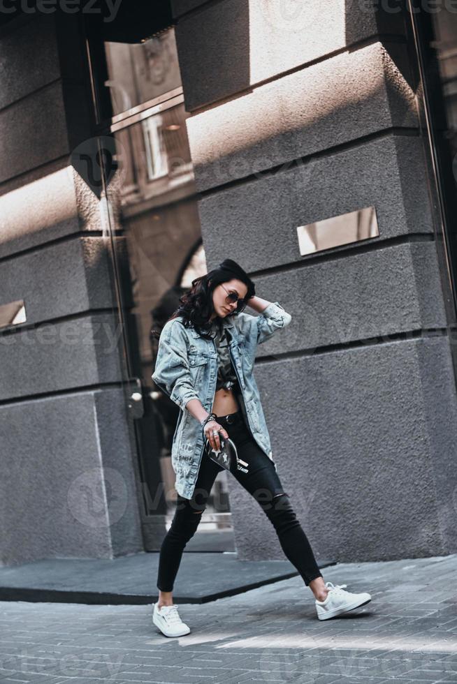 Charming girl on the move. Full length of beautiful young woman in denim jacket keeping hand in hair while walking down the street photo