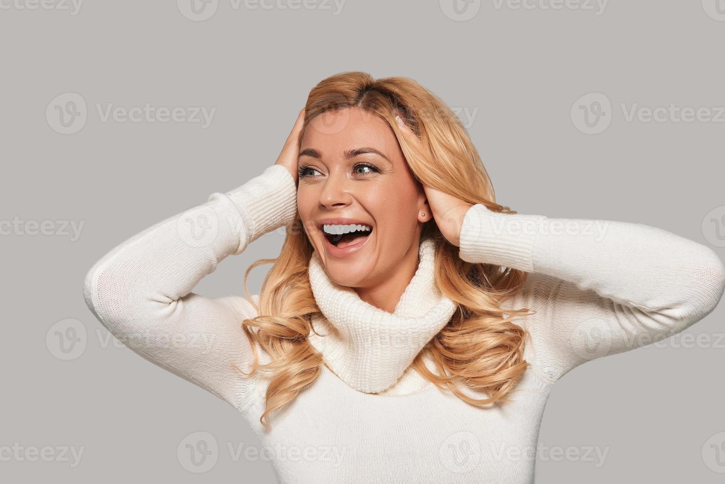 Extremely beautiful.  Attractive young smiling woman looking away and keeping hands in hair while standing against grey background photo