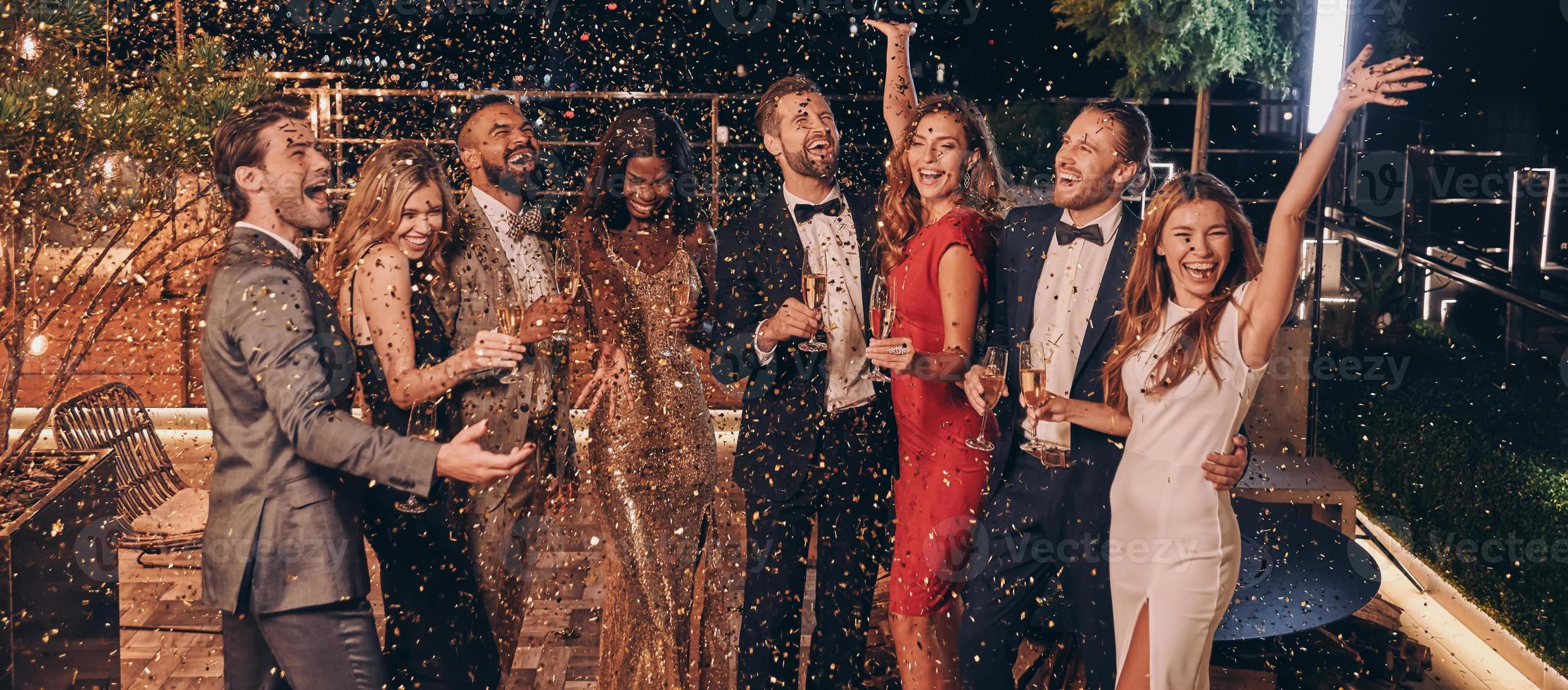 Group of happy people in formalwear having fun together with confetti flying all around photo