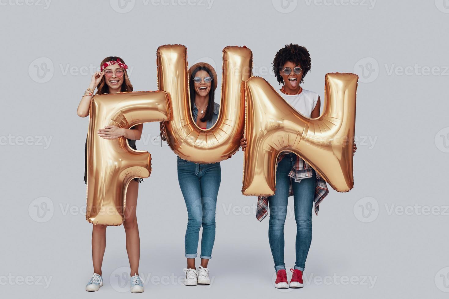 Full length of three attractive young women carrying balloons and smiling while standing against grey background photo