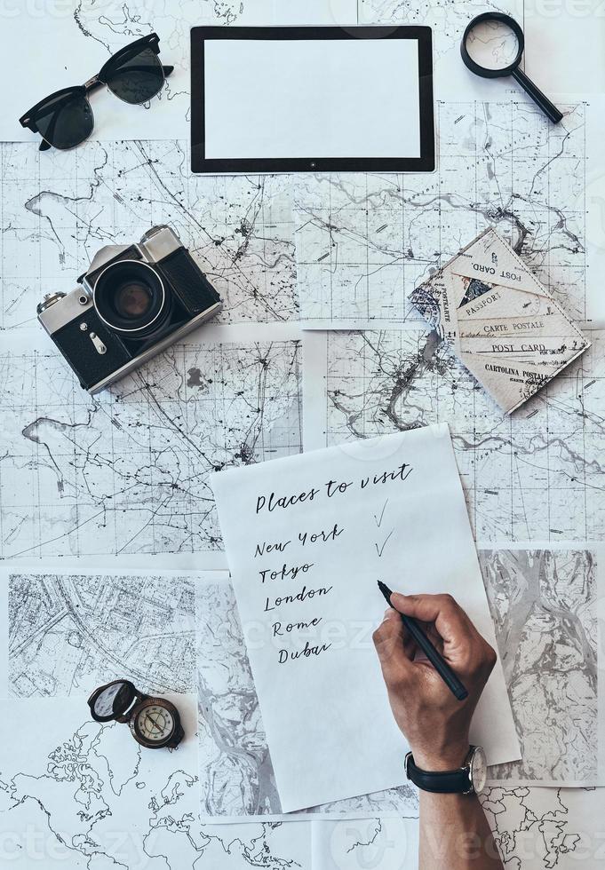 Places I want to visit. Close up top view of man writing down checklist with sunglasses, photo camera, compass, magnifying glass and passport lying on map around