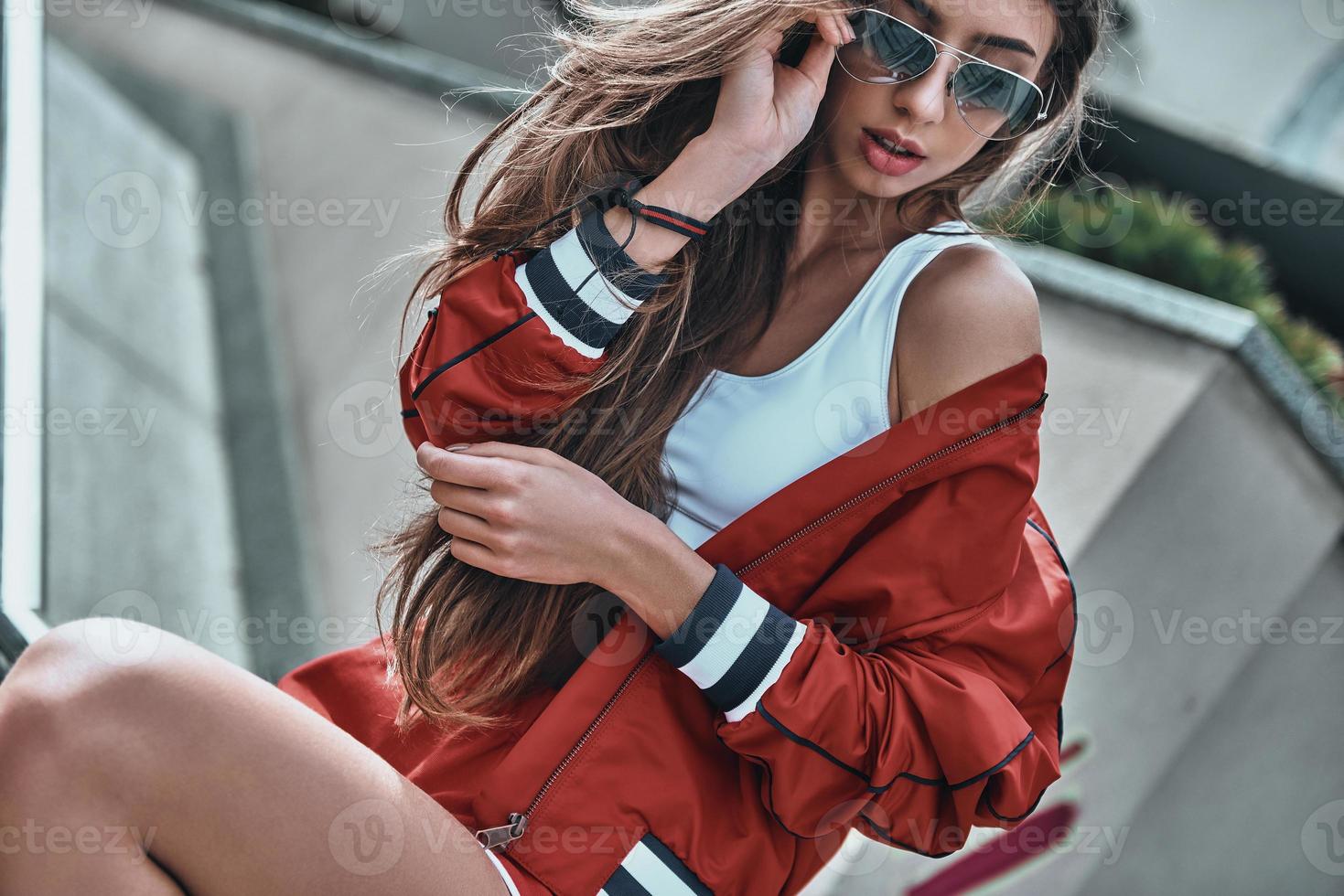 joven y genial. atractiva mujer joven con ropa deportiva ajustando sus gafas de sol mientras se sienta al aire libre foto