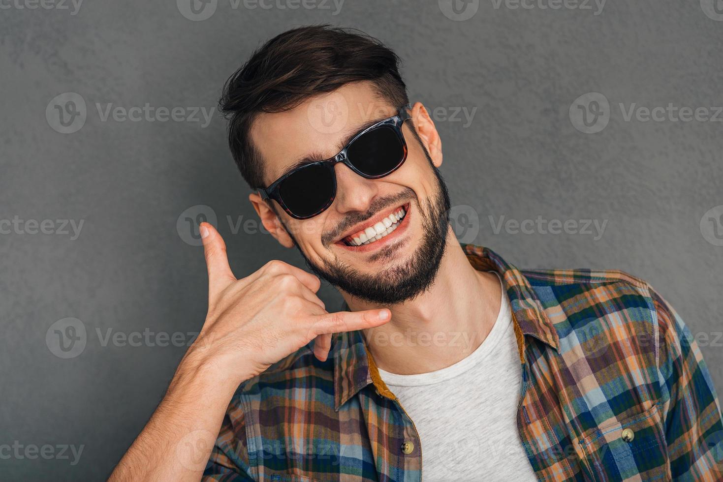 Call me Portrait of cheerful young man gesturing telephone with his hand and looking at camera with smile while standing against grey background photo