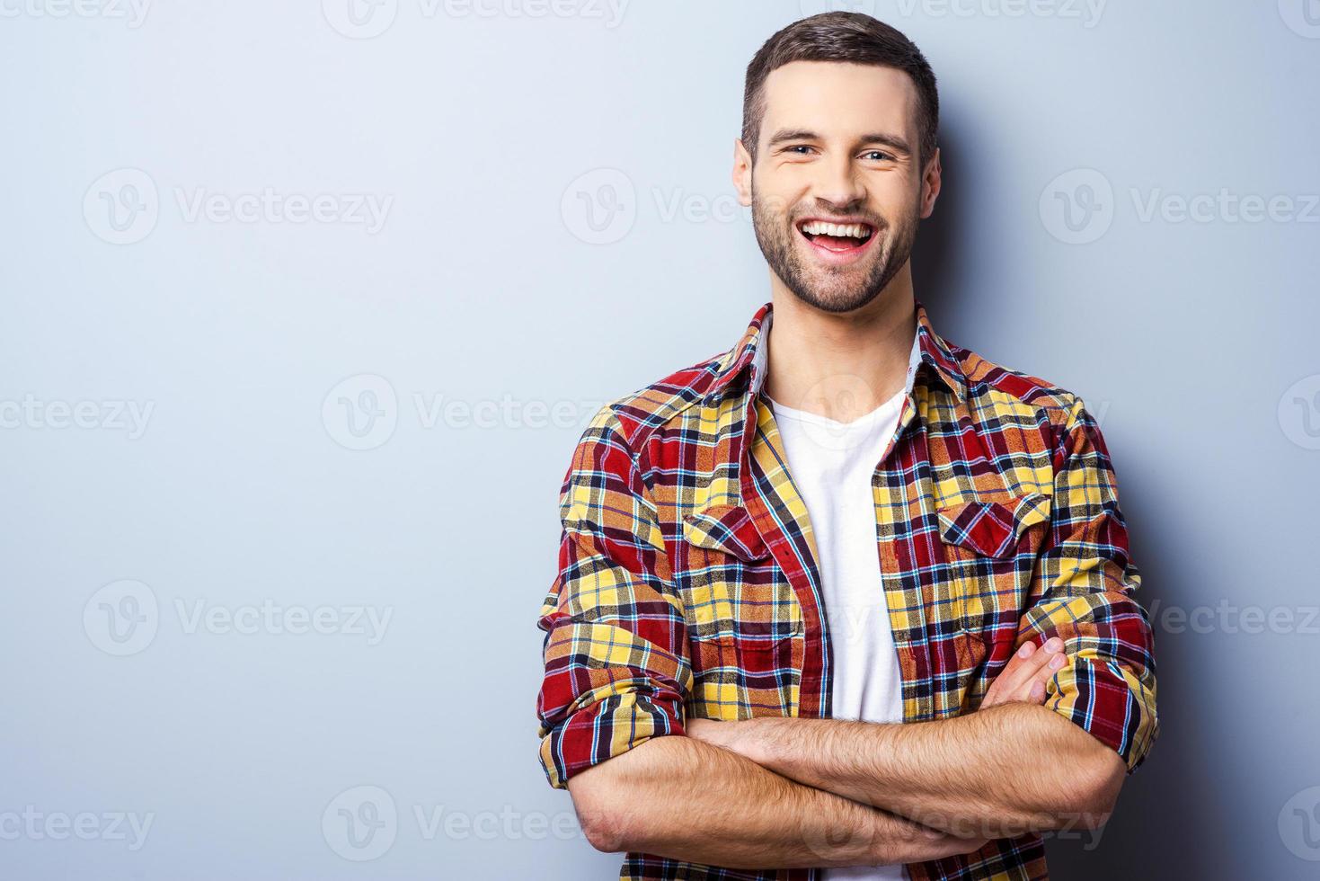 Happy young man. Portrait of handsome young man in casual shirt keeping arms crossed and smiling while standing against grey background photo