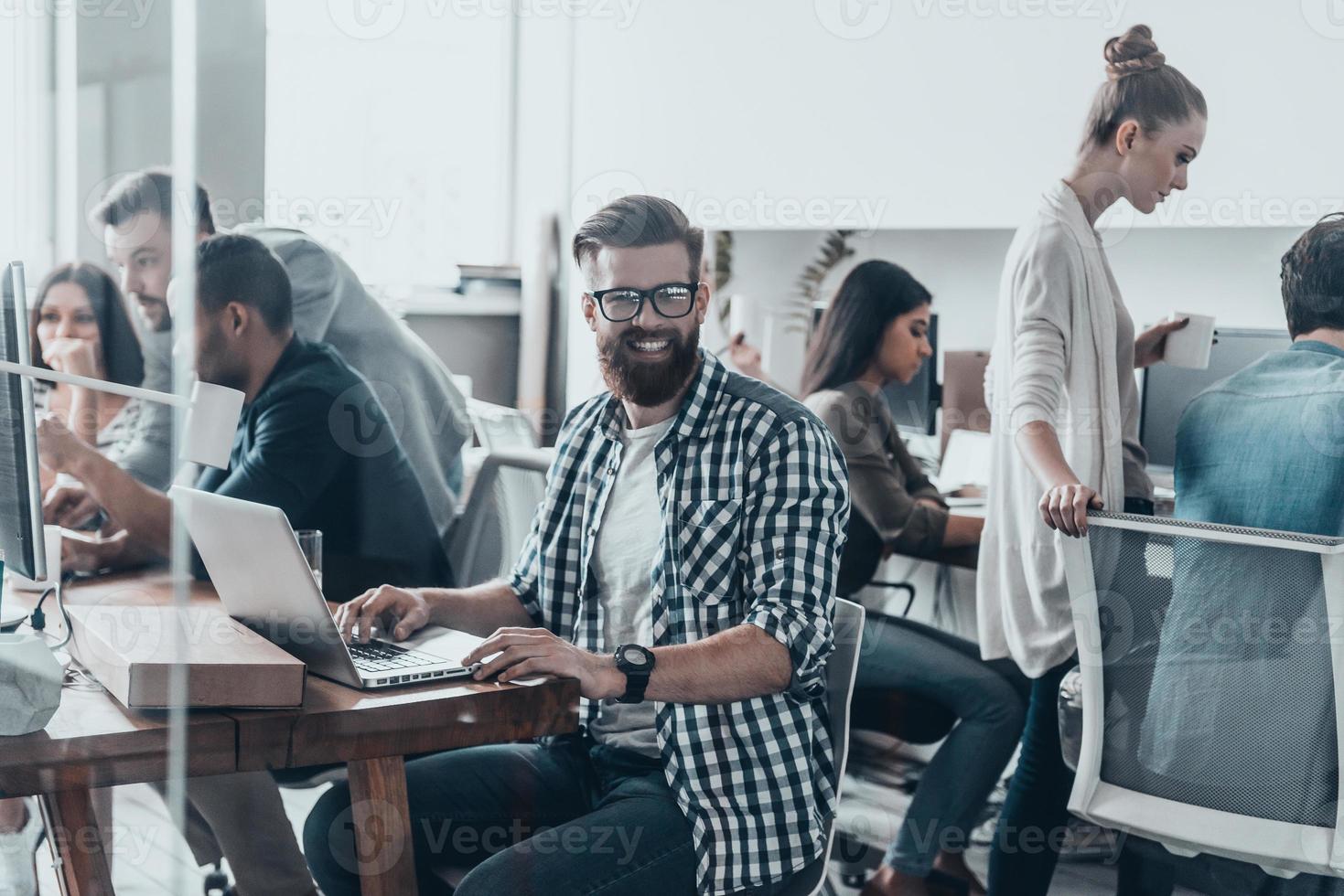 feliz de ser parte de un gran equipo. joven apuesto con barba sonriendo y mirando la cámara mientras se sienta en la oficina con colegas que trabajan en segundo plano foto