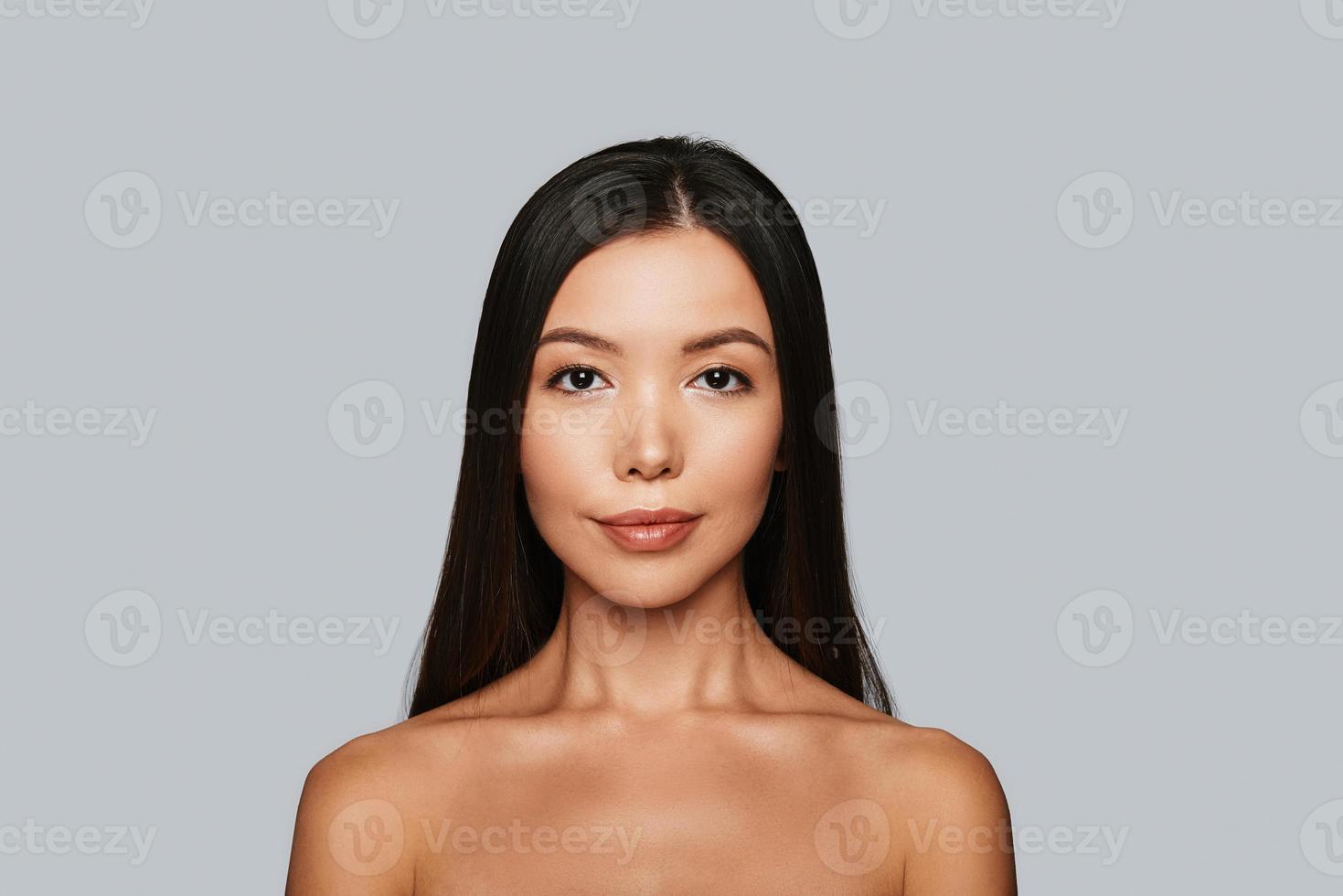 Glowing with natural beauty. Beautiful young Asian woman looking at camera and smiling while standing against grey background photo