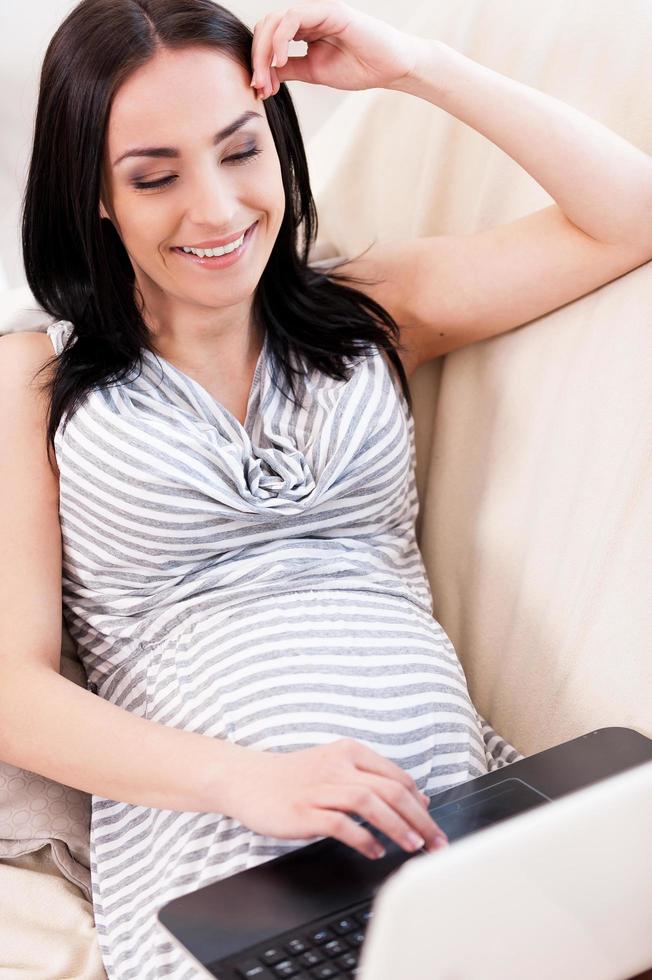 Reading about pregnancy. Top view of beautiful pregnant woman working on laptop and smiling while lying on a couch photo