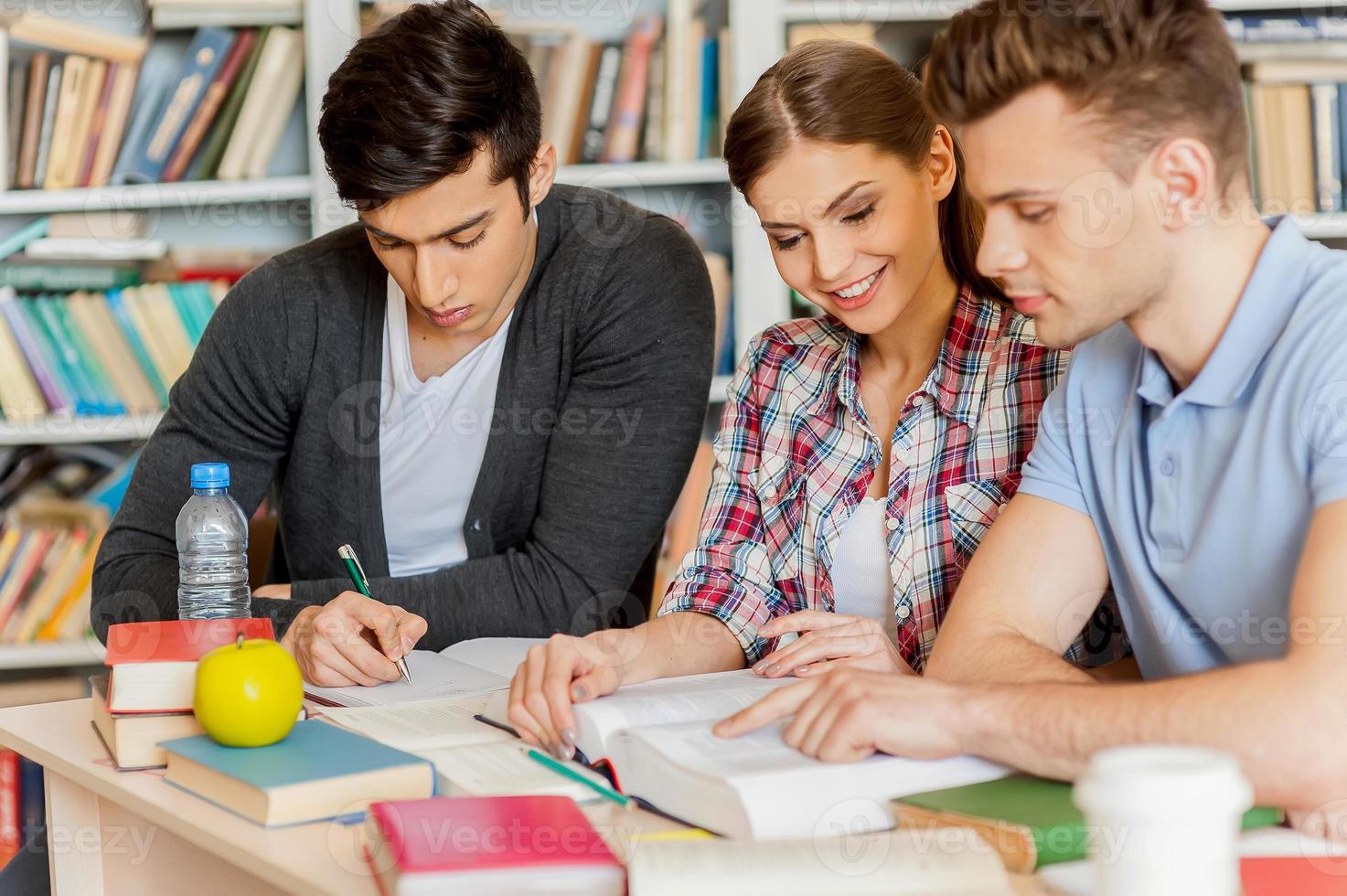 Preparing for exams in library. Three confident students reading a book together while sitting at the desk and against bookshelf in a library photo