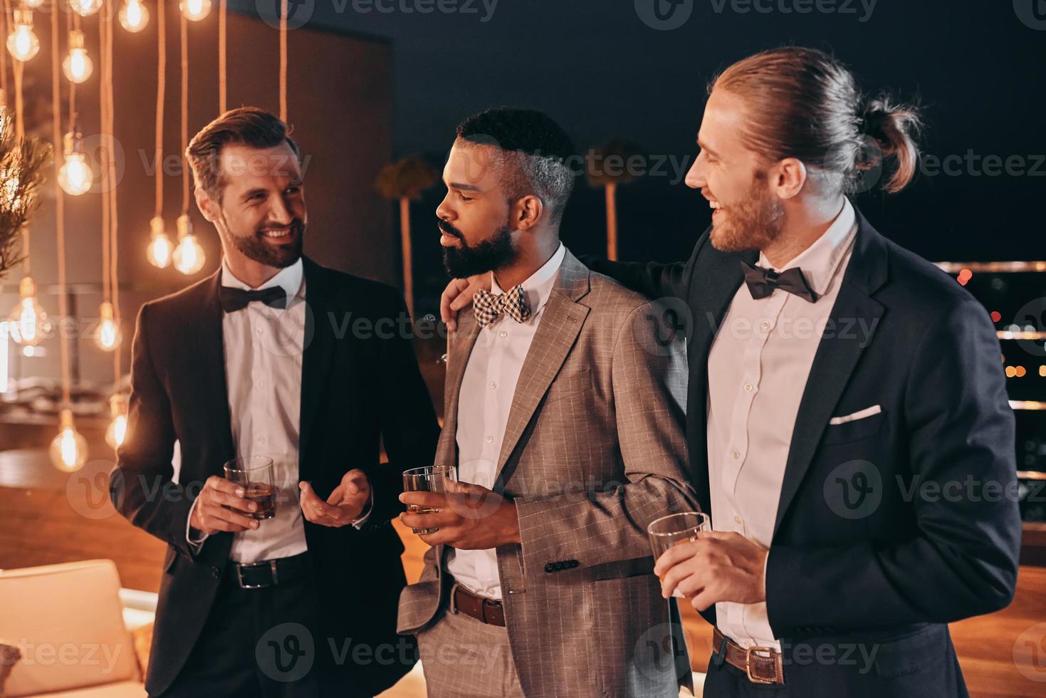 Three happy men in suits holding glasses with whiskey while spending time on party photo