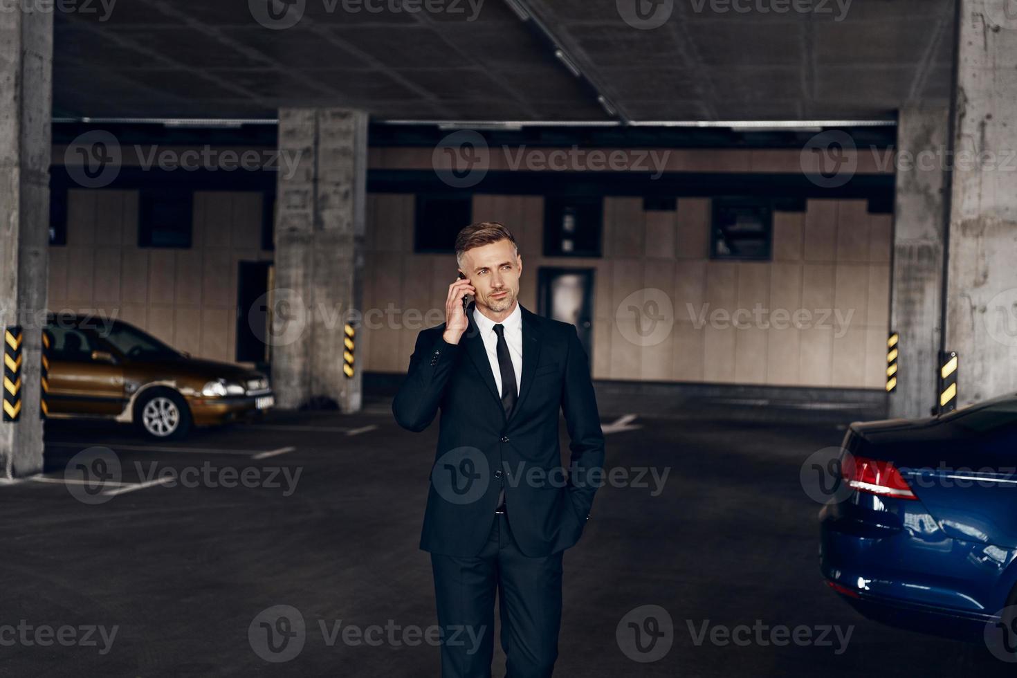 Confident mature businessman talking on mobile phone while walking by parking lot photo