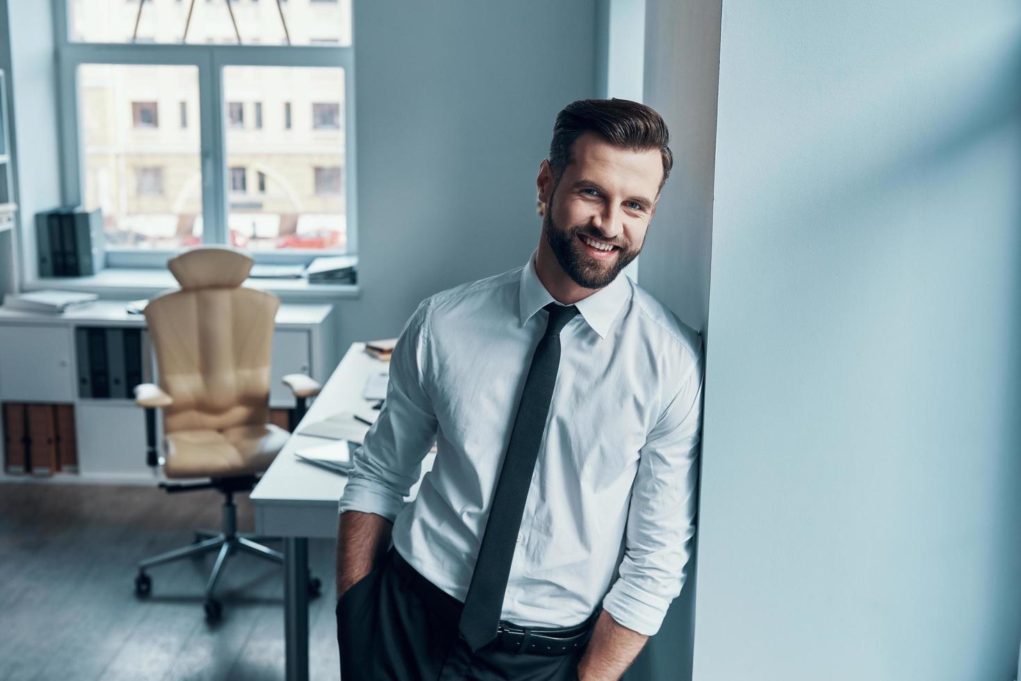 elegante joven con camisa y corbata mirando a la cámara y sonriendo mientras está de pie en la oficina foto