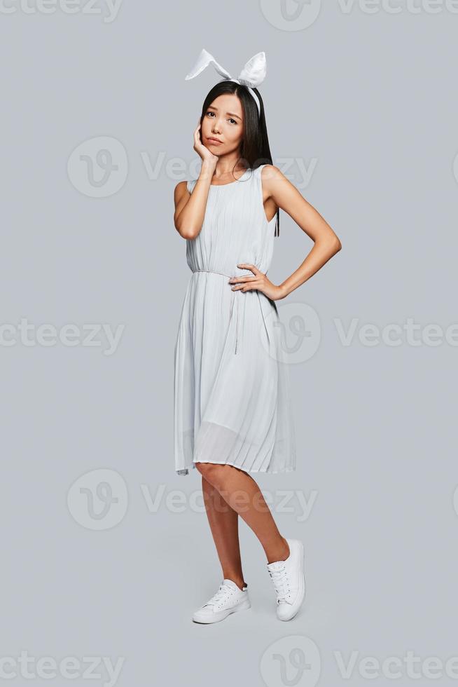So tired. Full length of young Asian woman in bunny ears looking at camera and making pity face while standing against grey background photo