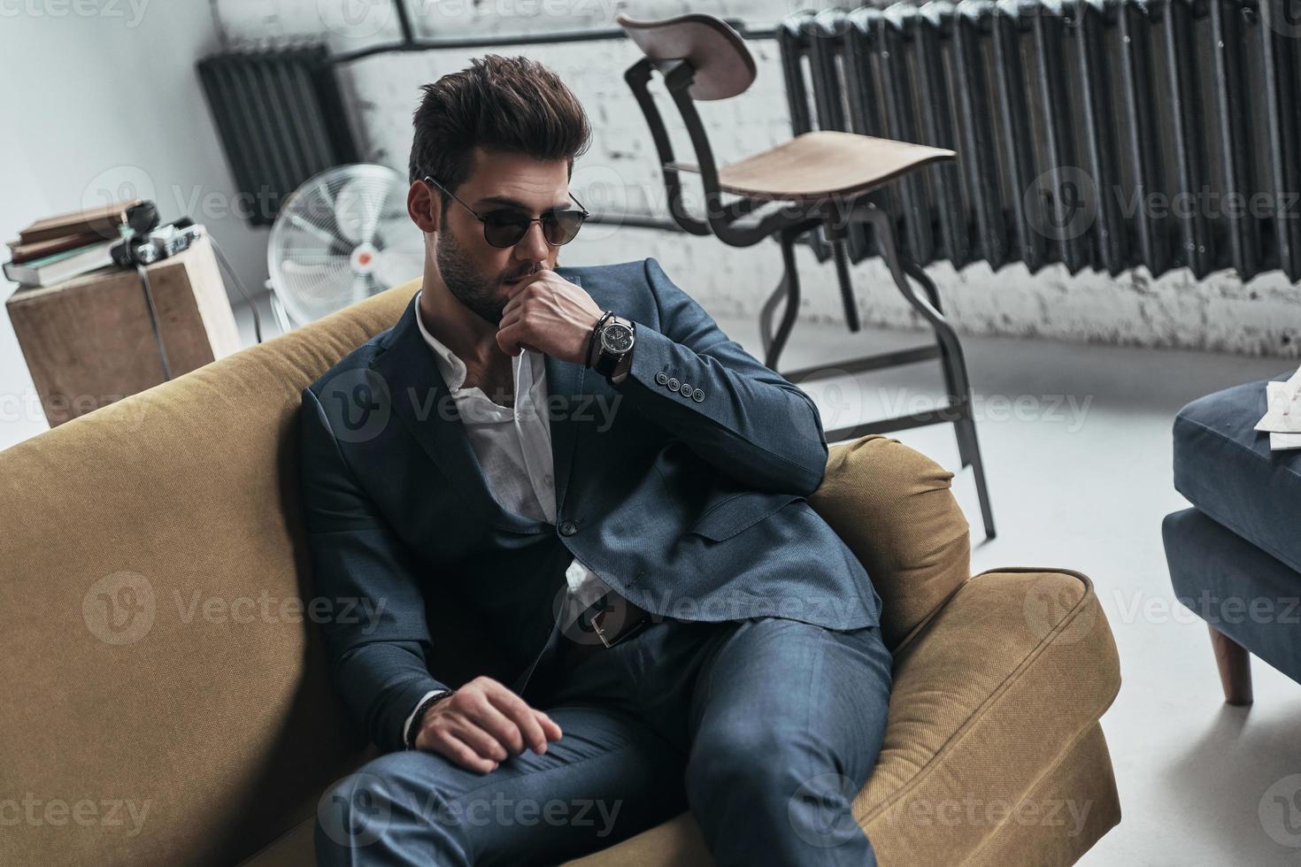 Confident in his perfect style. Thoughtful young man in full suit keeping hand on chin while sitting on the sofa at home photo