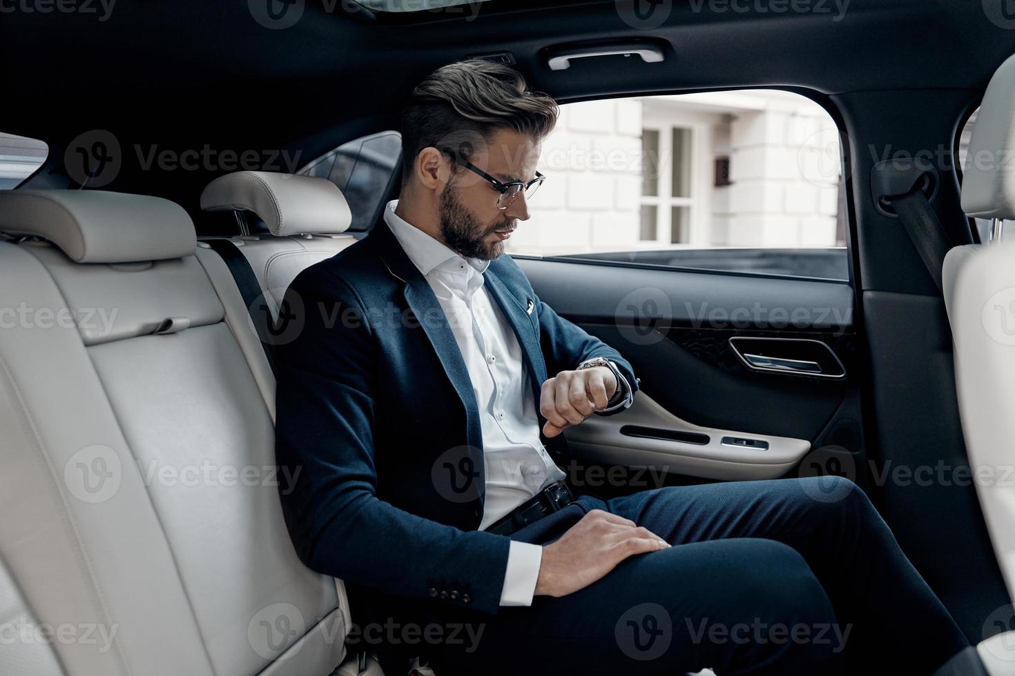 Keeping eye on time. Handsome young man in full suit looking at his watch while sitting in the car photo