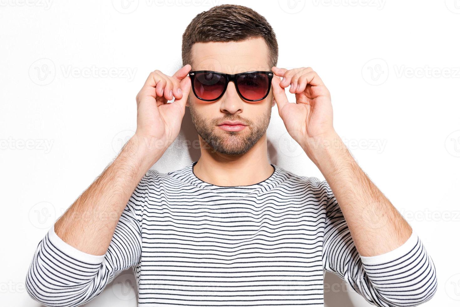 guapo y con estilo. apuesto joven ajustando sus gafas de sol y mirando a la cámara mientras está de pie contra el fondo blanco foto