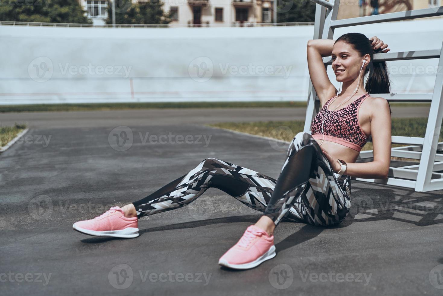 Minute to rest. Beautiful young woman in sports clothing smiling and looking away while sitting outdoors photo