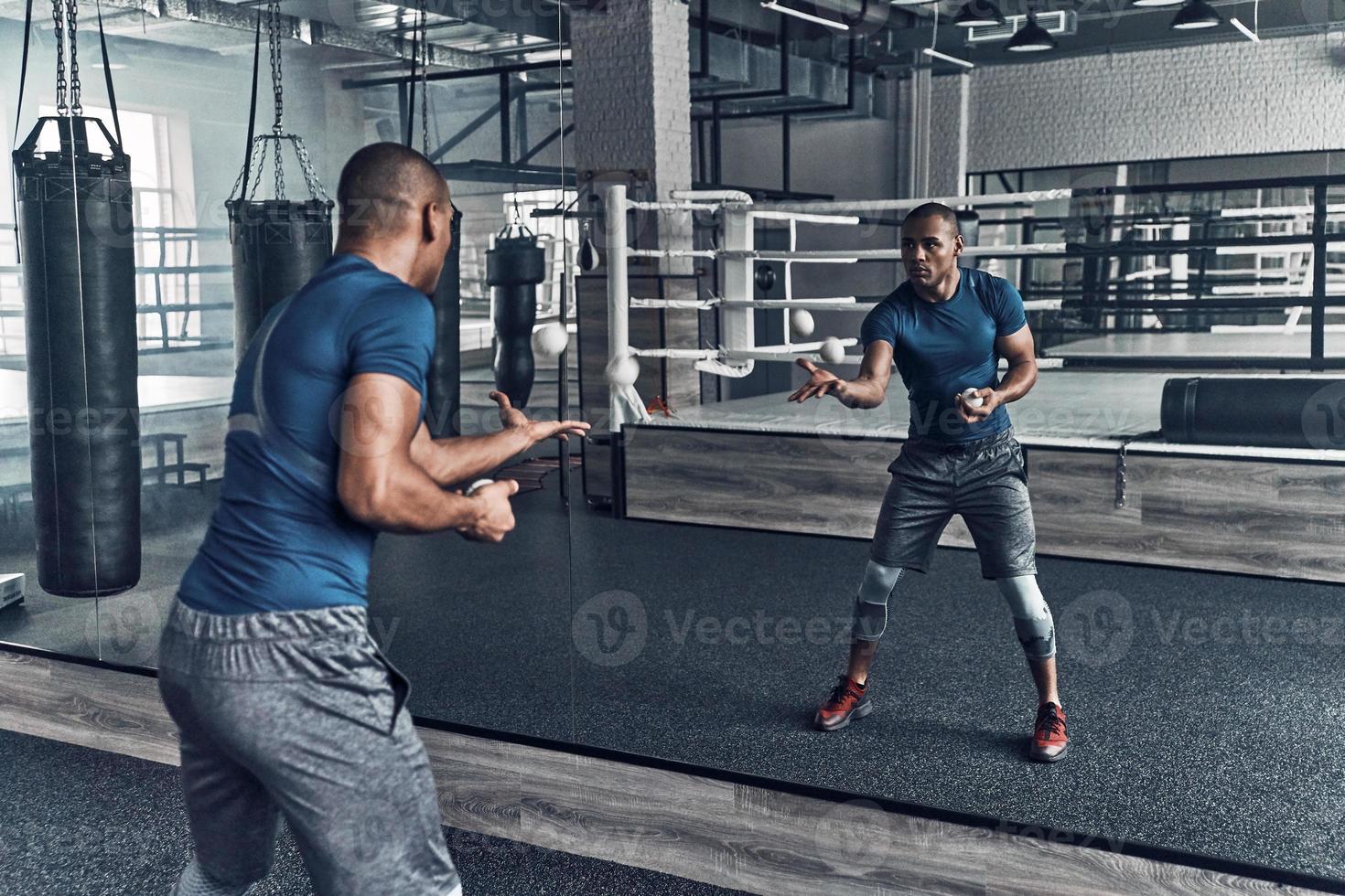 On the way to success. Handsome young African man in sport clothing exercising using balls while standing in front of the mirror in the gym photo