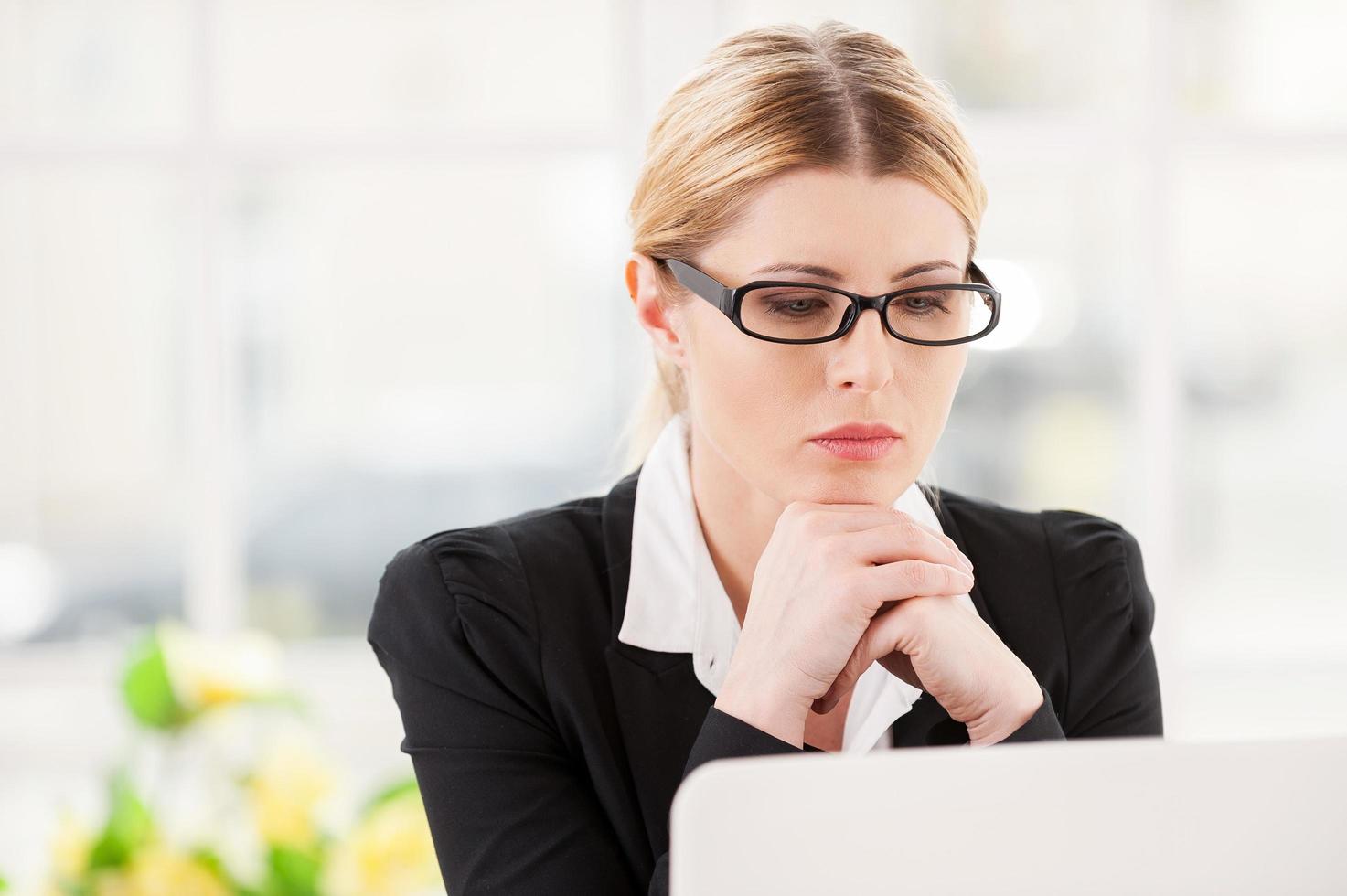 Concentrating at work. Serious mature woman in formalwear holding hands on chin and looking at laptop while sitting at her working place photo